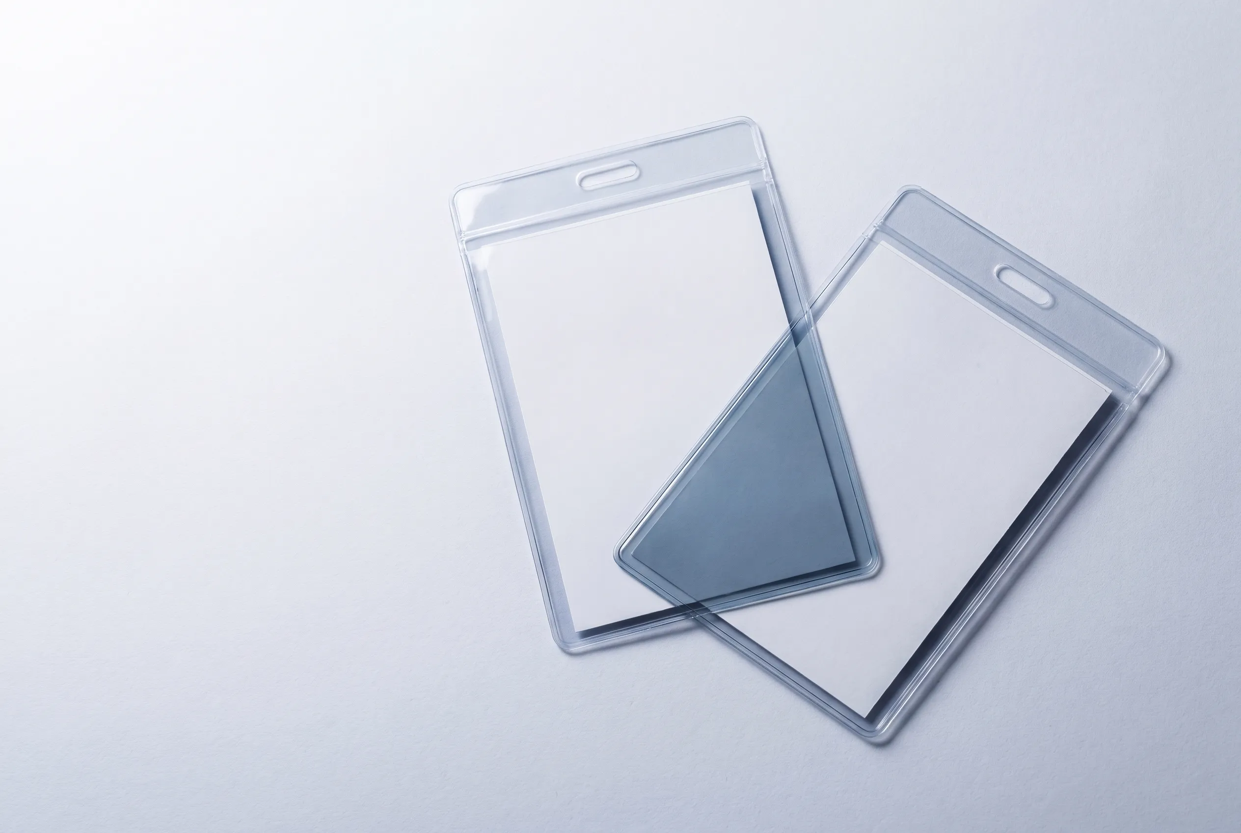 Two clear PVC lanyard pouches overlapping on a pale surface, the plastic deepening in tone where the layers cross.